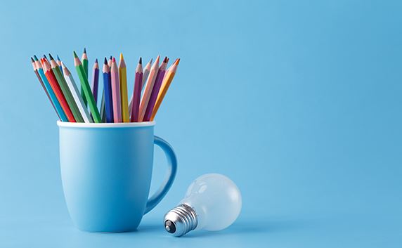 mug filled with color pencils and a light bulb next to it solid blue background